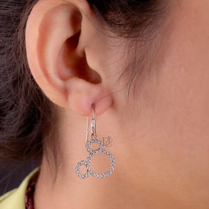 May include: Silver-toned dangle earrings featuring a Mickey Mouse-inspired design. Each earring has three connected circles, outlined with small, sparkling stones. The earrings are suspended from a simple hook.