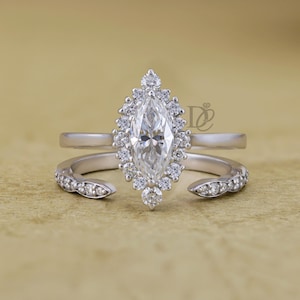 May include: A silver-coloured ring featuring a central marquise-cut stone surrounded by a halo of small round stones. The band is open at the front, with additional small round stones set along the sides of the open ends. A logo is visible.
