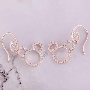May include: A pair of rose gold earrings in the shape of a mouse head, encrusted with small, sparkling clear crystals. The earrings have a hook closure and are displayed on a light-colored surface.