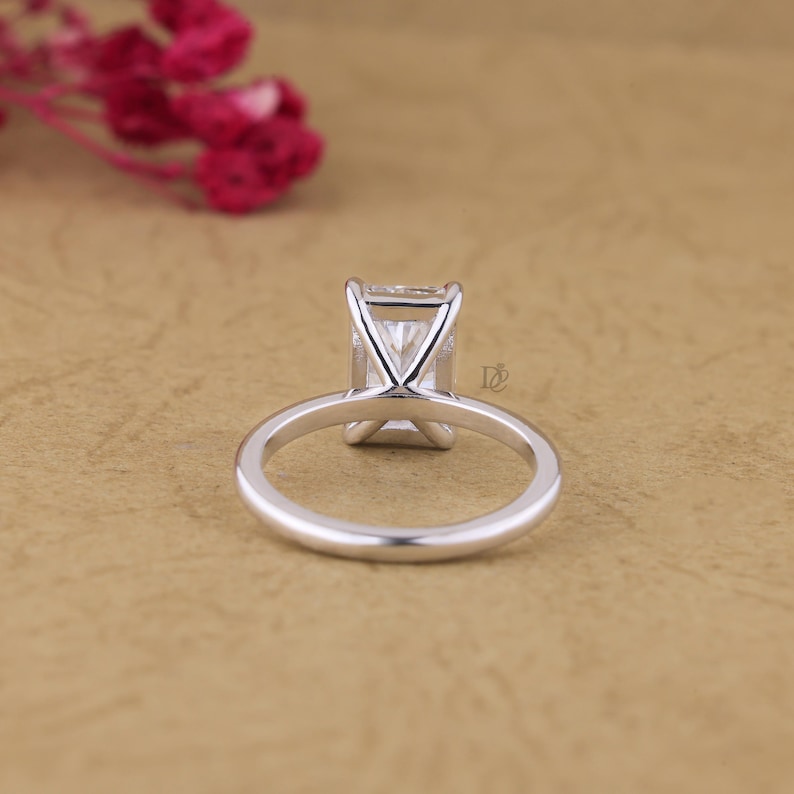 May include: A silver-coloured ring featuring a rectangular-cut clear gemstone set in a four-prong basket setting. The band is smooth and polished, showcasing a clean, metallic finish. A small logo is visible on the side of the setting.