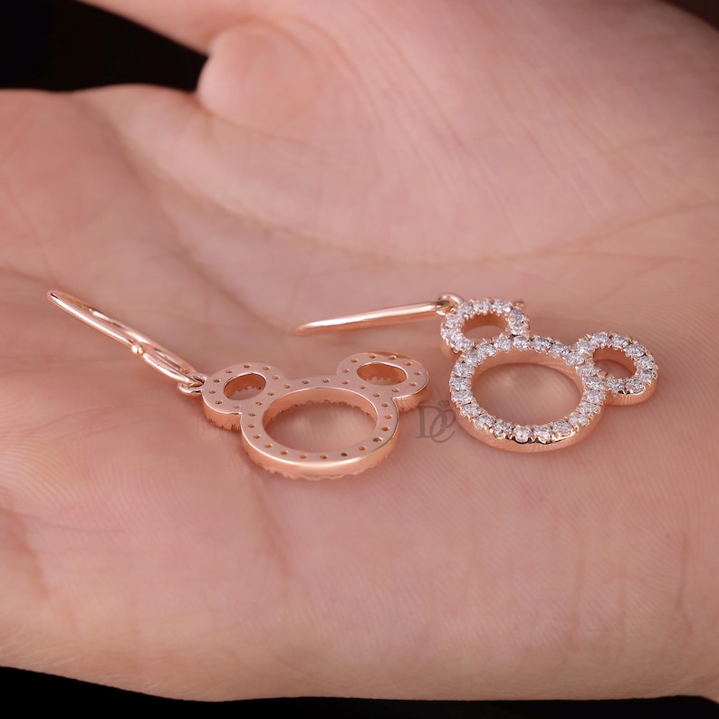 May include: Rose gold earrings in the shape of Mickey Mouse's head. The earrings are encrusted with small, sparkling white stones. The design features a classic three-circle silhouette, with a hook for pierced ears.