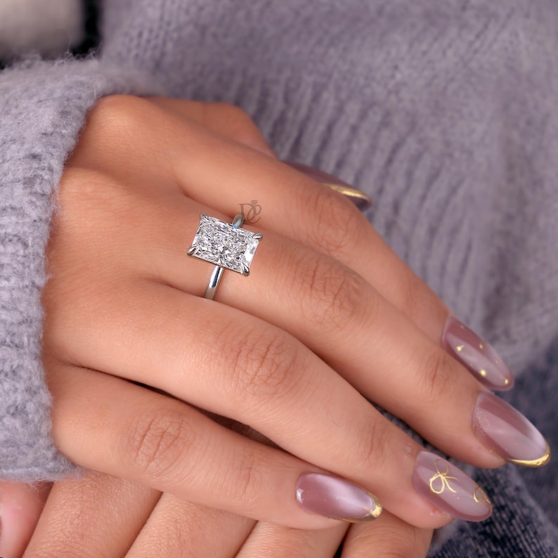 May include: A silver-toned ring featuring a large, rectangular, brilliant-cut clear stone set in a four-prong setting. The band is thin and polished, showcasing a clear, faceted gemstone that reflects light across its surface.