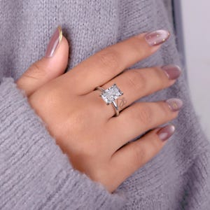 May include: A silver-toned ring featuring a large, rectangular, faceted clear stone set in a four-prong setting. The band is thin and polished, showcasing a clean, metallic finish that complements the central stone's brilliant, multi-faceted cut.