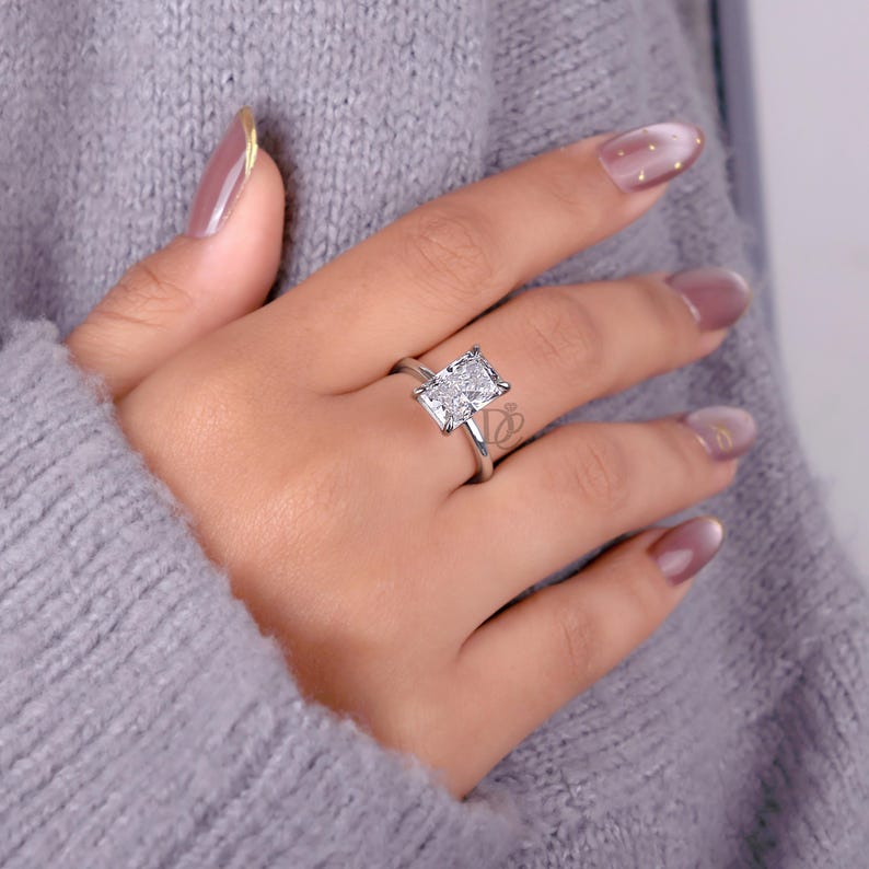 May include: A silver-toned ring featuring a large, rectangular, faceted clear stone set in a four-prong setting. The band is thin and polished, showcasing a clean, metallic finish that complements the central stone's brilliant, multi-faceted cut.