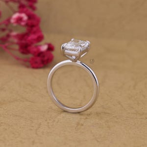 May include: A silver-toned ring featuring a rectangular radiant-cut clear gemstone held in a four-prong setting. The band is smooth and polished, showcasing a clean, minimalist design. A small logo is engraved on the side of the metal setting.