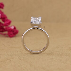 May include: A silver-toned ring featuring a single rectangular-cut clear gemstone set in a four-prong setting. The band is smooth and circular, and a small logo consisting of the letters DE is visible on the side of the metal setting.