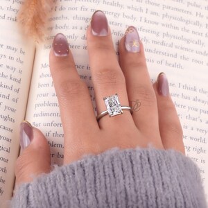 May include: A silver-toned ring featuring a large, rectangular, clear faceted gemstone set in a four-prong setting. The ring is worn on a finger, positioned against an open book with visible printed text, showcasing the clear stone's brilliance.