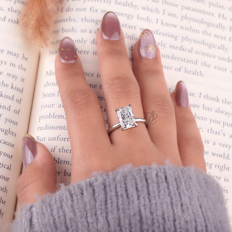 May include: A silver-toned ring featuring a large, rectangular, clear faceted gemstone set in a four-prong setting. The ring is worn on a finger, positioned against an open book with visible printed text, showcasing the clear stone's brilliance.