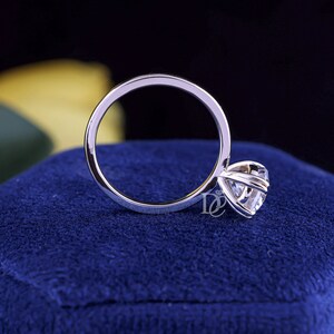 May include: A silver-toned metal ring featuring a single round-cut clear gemstone held in a six-prong setting. The band is smooth and circular. A small logo consisting of the letters DC with a diamond icon is visible near the gemstone setting.