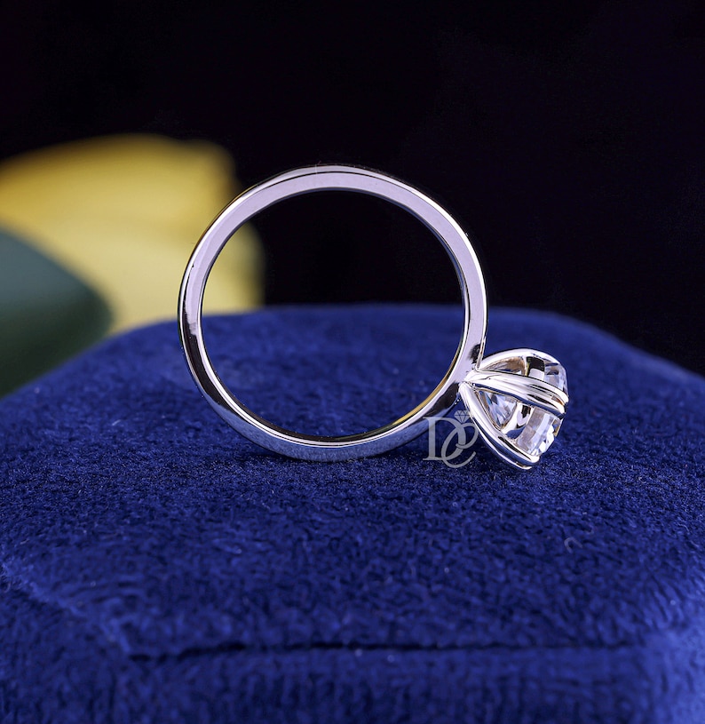 May include: A silver-toned metal ring featuring a single round-cut clear gemstone held in a six-prong setting. The band is smooth and circular. A small logo consisting of the letters DC with a diamond icon is visible near the gemstone setting.