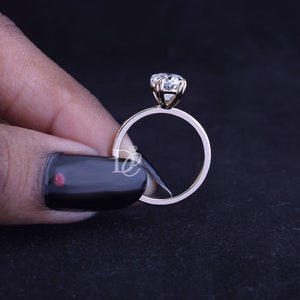 May include: A gold ring featuring a single round-cut clear gemstone held in a six-prong setting. The band is thin and polished, showcasing a minimalist design. A small white logo with the letters DC and a diamond icon is visible on the band.