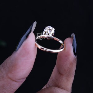 May include: A gold ring featuring a single oval-cut clear gemstone held in a multi-prong setting. The band is smooth and polished, showcasing a reflective metallic finish. A small logo with the letters DJ is visible near the stone setting.