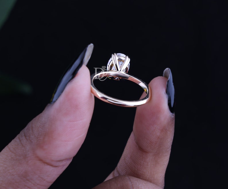 May include: A gold ring featuring a single oval-cut clear gemstone held in a multi-prong setting. The band is smooth and polished, showcasing a reflective metallic finish. A small logo with the letters DJ is visible near the stone setting.