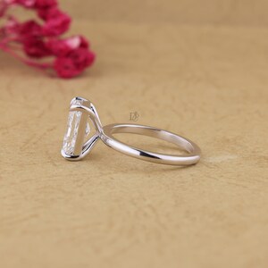 May include: A silver-coloured ring featuring a large, rectangular, clear faceted gemstone set in a high-profile prong setting. The band is smooth and polished, with a small logo mark visible on the side of the setting near the stone.