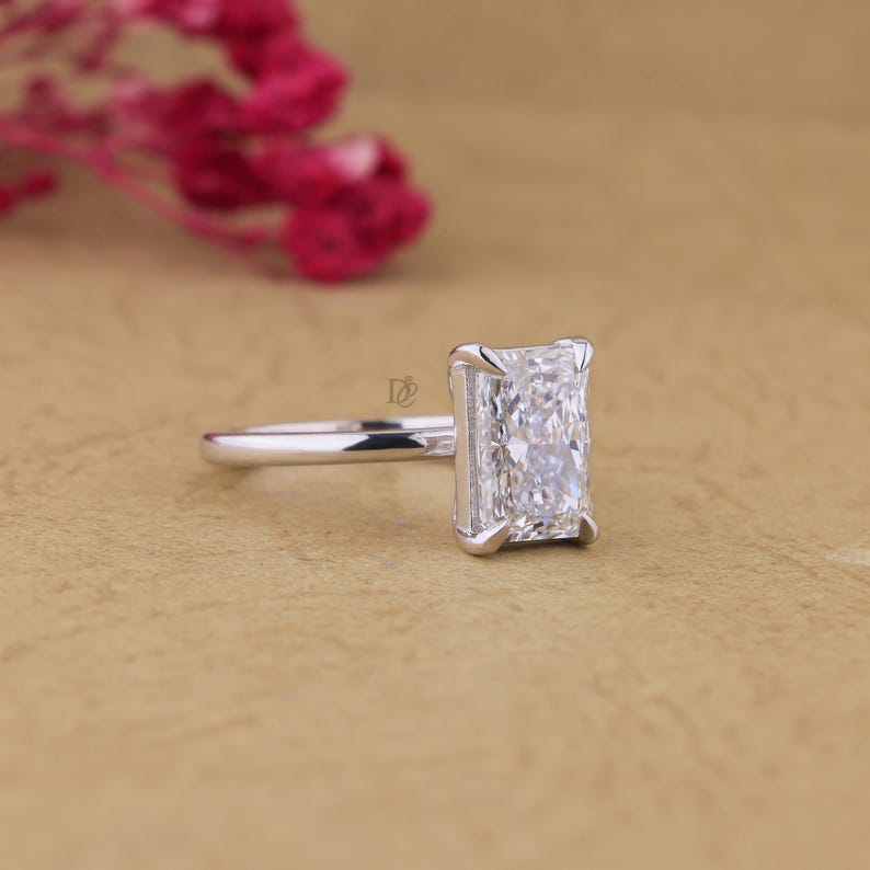 May include: A silver-coloured ring featuring a rectangular radiant-cut clear gemstone set in a four-prong setting. The band is smooth and polished, showcasing a clean design. A small logo is visible on the side of the setting near the gemstone.