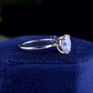 May include: A silver-toned ring featuring a single round, clear, faceted gemstone set in a four-prong setting. The band is smooth and polished. A small logo with the letters DB and a diamond icon is visible near the gemstone setting.