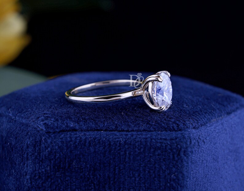 May include: A silver-toned ring featuring a single round, clear, faceted gemstone set in a four-prong setting. The band is smooth and polished. A small logo with the letters DB and a diamond icon is visible near the gemstone setting.