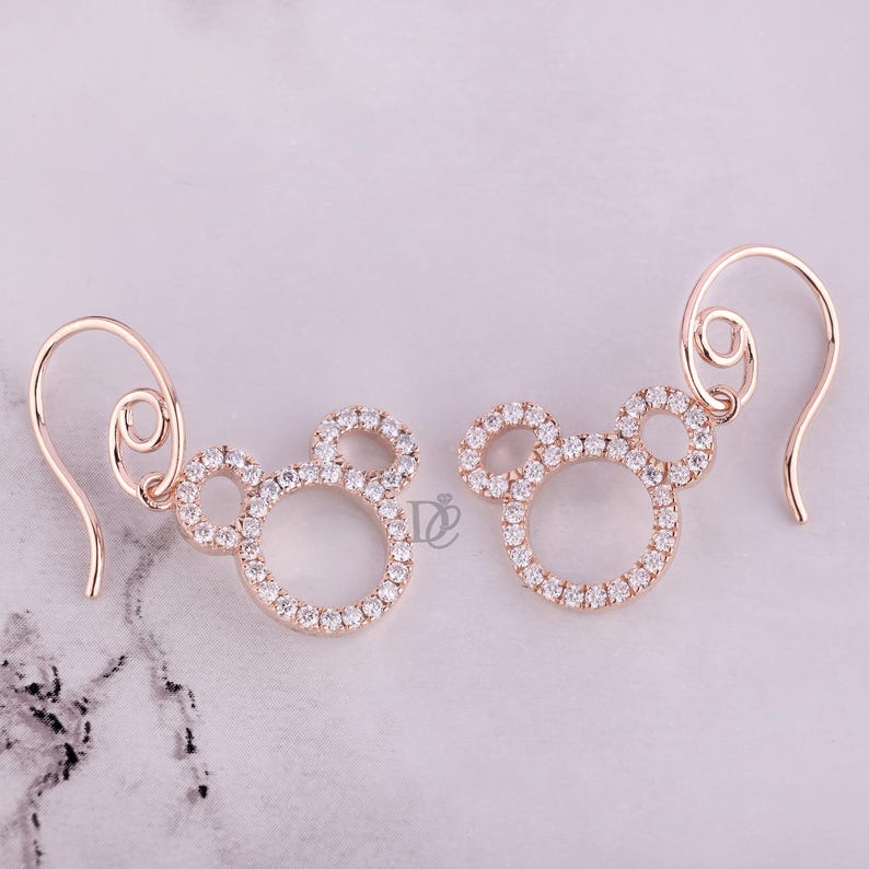 May include: A pair of rose gold earrings in the shape of a mouse head, encrusted with small, sparkling clear crystals. The earrings have a hook closure and are displayed on a light-colored surface.