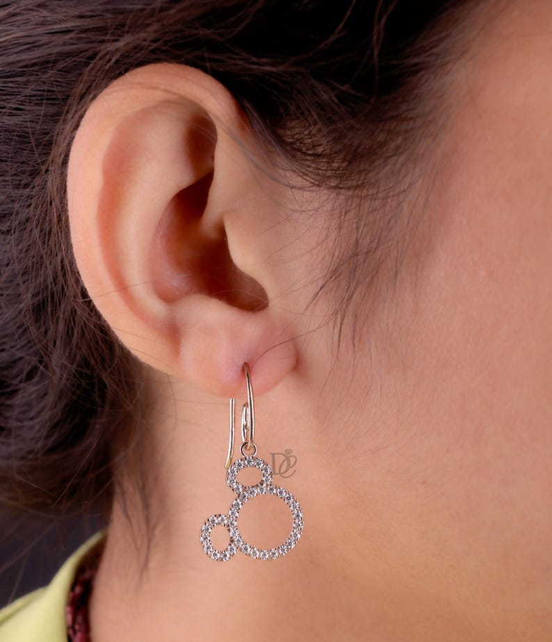 May include: Silver-toned dangle earrings featuring a Mickey Mouse-inspired design. Each earring has three connected circles, outlined with small, sparkling stones. The earrings are suspended from a simple hook.