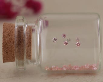 May include: A clear glass bottle with a cork stopper containing numerous small, round, faceted pink gemstones. Four of the pink stones are positioned against the side of the glass, while the rest are gathered at the bottom of the bottle.