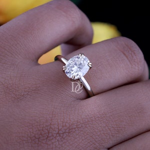 May include: A gold ring featuring a large, oval-cut, clear, faceted gemstone held in place by four prongs. The band is smooth and polished, and a small logo with the letters DC and a diamond icon is visible on the side of the ring setting.