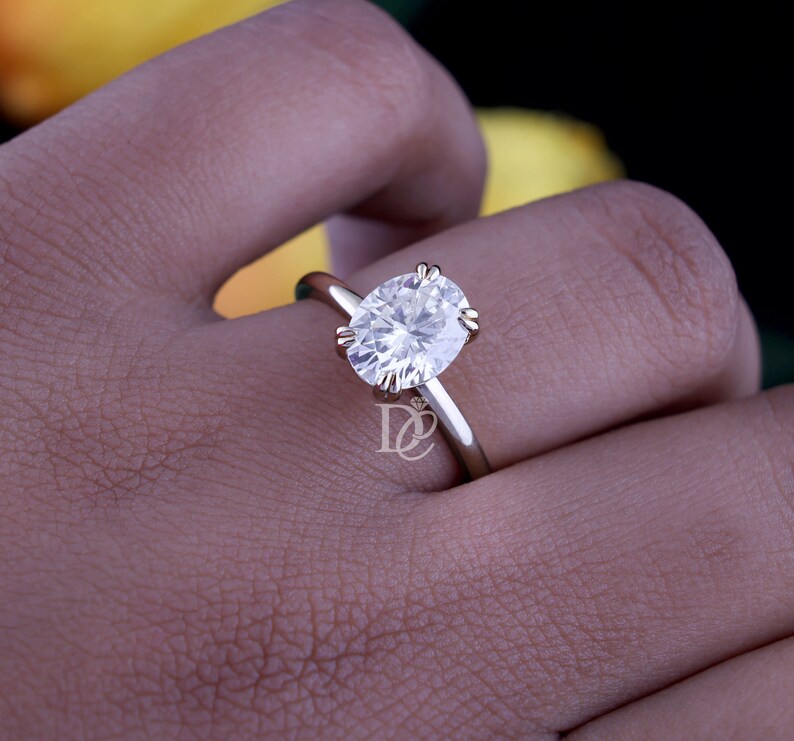 May include: A gold ring featuring a large, oval-cut, clear, faceted gemstone held in place by four prongs. The band is smooth and polished, and a small logo with the letters DC and a diamond icon is visible on the side of the ring setting.