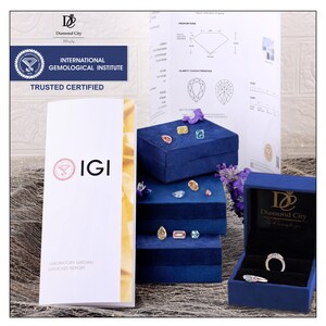 Op de afbeelding: Een verzameling losse gekleurde diamanten en twee ringen op gestapelde blauwe fluwelen doosjes. Een IGI-laboratoriumrapport voor in het lab gekweekte diamanten en een Diamond City-certificaat zijn zichtbaar, met details over proporties en zuiverheid.