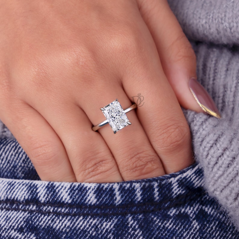 May include: A silver-coloured ring featuring a large, rectangular radiant-cut clear gemstone set in a four-prong setting on a thin, polished metal band. The ring is positioned on a finger, showcasing the clear, faceted stone and the metallic band.