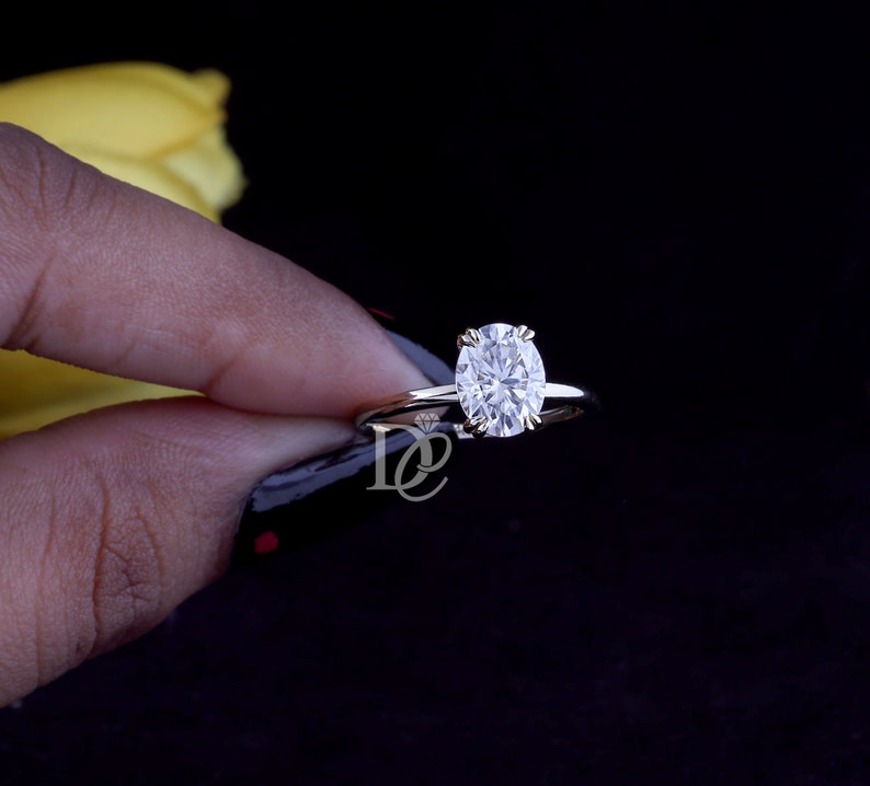 May include: A silver-toned ring featuring a single oval-cut clear gemstone set in a four-prong setting. The band is thin and polished, and a small logo with the letters DE and a diamond icon is visible on the metal surface near the setting.