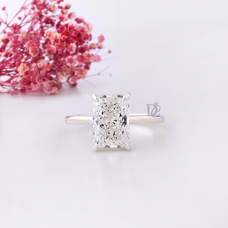 May include: A radiant-cut diamond engagement ring with a slender silver band. The large, rectangular diamond sparkles, reflecting light. Pink flowers are softly blurred in the background, adding a touch of color and elegance. A classic and timeless piece of jewelry.