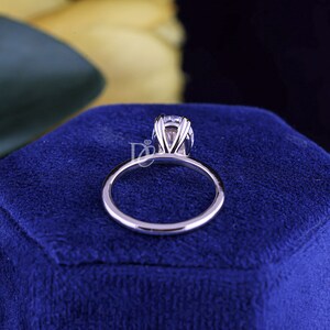 May include: A silver-toned ring featuring a single round faceted clear gemstone held in a multi-prong setting. The band is smooth and polished, showcasing a metallic luster. A small logo with the letters DC and a diamond icon is visible.