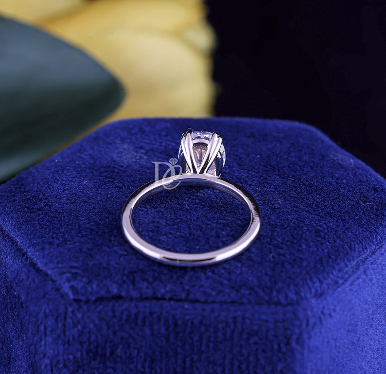 May include: A silver-toned ring featuring a single round faceted clear gemstone held in a multi-prong setting. The band is smooth and polished, showcasing a metallic luster. A small logo with the letters DC and a diamond icon is visible.