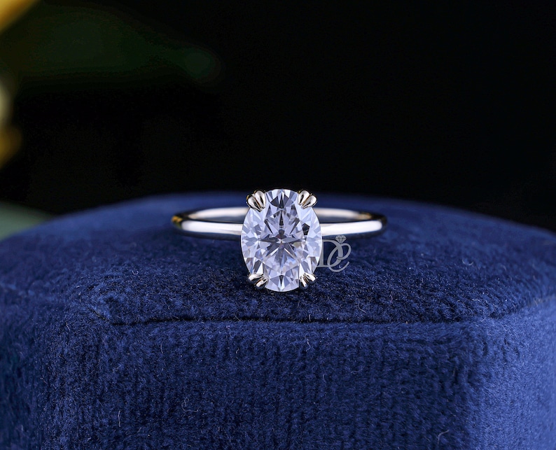 May include: A silver-toned ring featuring a single oval-cut clear gemstone held by four prongs on a smooth, polished band. The ring is centered on a dark blue velvet surface and includes a small logo mark near the base of the gemstone setting.