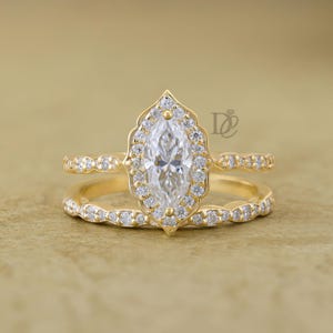 May include: A gold ring set featuring a central marquise-cut stone surrounded by a halo of small round stones. The setting has a scalloped edge, and the band is adorned with a row of small round stones. The letters DE are visible.