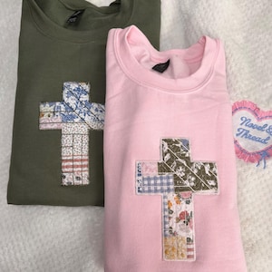 May include: An olive green sweatshirt and a light pink sweatshirt, each featuring a large cross applique made of various floral and plaid fabric patches. A heart-shaped patch with the text Novel & Thread is visible to the right.