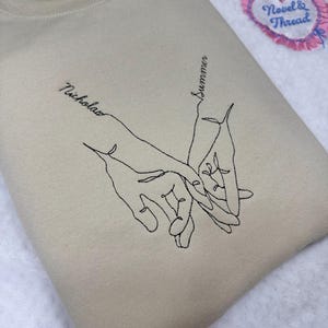May include: Beige sweatshirt featuring an embroidered line art design of two hands holding each other. The names "Nicholas" and "Summer" are embroidered on the sweatshirt. A pink heart-shaped patch with the text "Novel & Thread" is in the background.