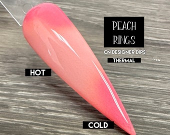 May include: A thermal nail dip powder in a peach color that changes to a pink color when cold. The text "Peach Rings" is written in black on a white background. The text "CN Designer Dips" is written in black on a white background. The text "Thermal" is written in black on a white background. The text "HOT" is written in black on a white background. The text "COLD" is written in black on a white background.