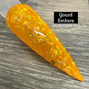 May include: A nail swatch with a vibrant orange hue, speckled with iridescent gold and green flakes. The text "Gourd Embers" and "CN DESIGNER DIPS" are displayed in a black rectangle. The nail is tapered and glossy.