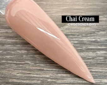 May include: A single nail dipped in a light brown color, with a glossy finish. The nail is labeled "Chai Cream" by CN Designer Dips. The nail is long and pointed, and is displayed against a wooden surface.