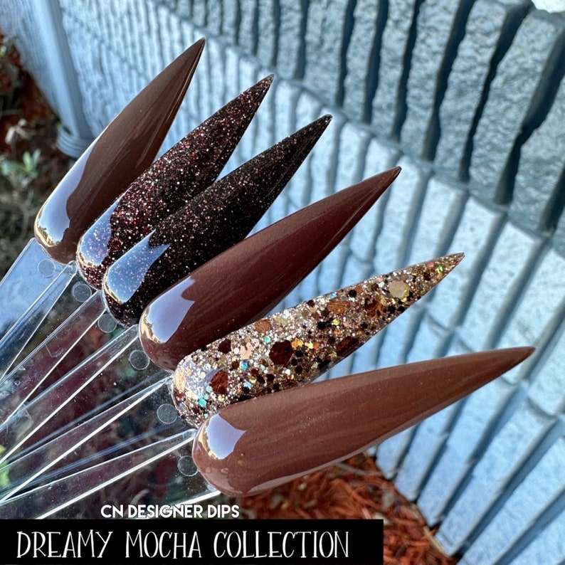 May include: Five different shades of brown nail dip powder, including a glitter option. The colors range from light to dark brown. The text "CN Designer Dips Dreamy Mocha Collection" is at the bottom of the image.