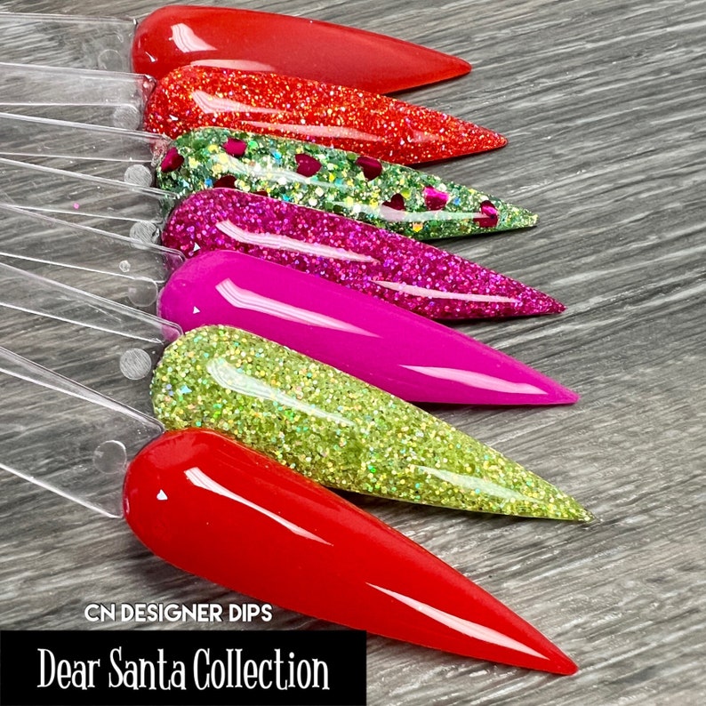 May include: Six different shades of nail dip powder in a variety of colors, including red, green, and pink. The dip powders are arranged in a fan shape on a wooden surface. The text "CN DESIGNER DIPS Dear Santa Collection" is visible at the bottom of the image.