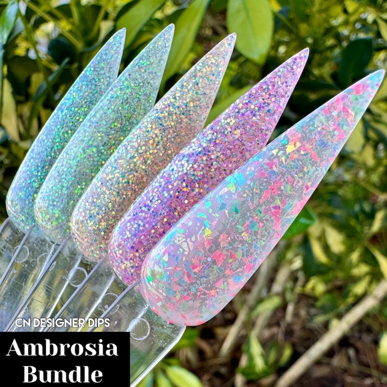 Puede incluir: Un conjunto de cuatro polvos para u&ntilde;as con purpurina iridiscente en una variedad de colores, incluyendo rosa, morado y blanco. Los polvos est&aacute;n dispuestos en una fila sobre una superficie de pl&aacute;stico transparente. El texto "Ambrosia Bundle" es visible en la superficie.