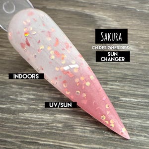 May include: A nail swatch with a gradient design, transitioning from clear to pink. The nail is adorned with glitter and the text "SAKURA", "CN DESIGNER DIPS", and "SUN CHANGER". The words "INDOORS" and "UV/SUN" are also visible.