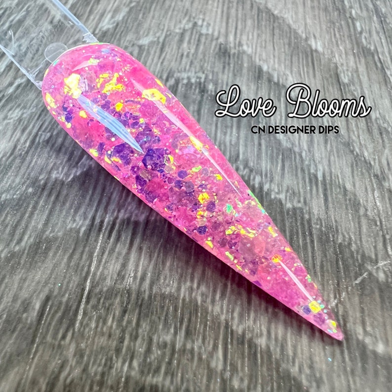 Op de afbeelding: Roze nageldippoeder met gouden en iriserende glitter. Het label luidt "Love Blooms CN Designer Dips."