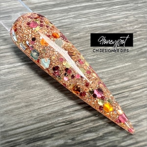 May include: A close-up of a long, pointed, clear nail with a glittery, multi-colored design. The nail is covered in a variety of small, iridescent glitter pieces, including gold, red, and orange. The nail is on a wooden surface. The text "Honeyfoot CN DESIGNER DIPS" is visible in the upper right corner of the image.