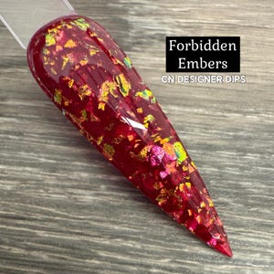 May include: A long, pointed nail sample with a deep red base and shimmering gold and iridescent flakes. The text "Forbidden Embers" and "CN DESIGNER DIPS" are displayed in a black box. The nail is on a gray wooden surface.