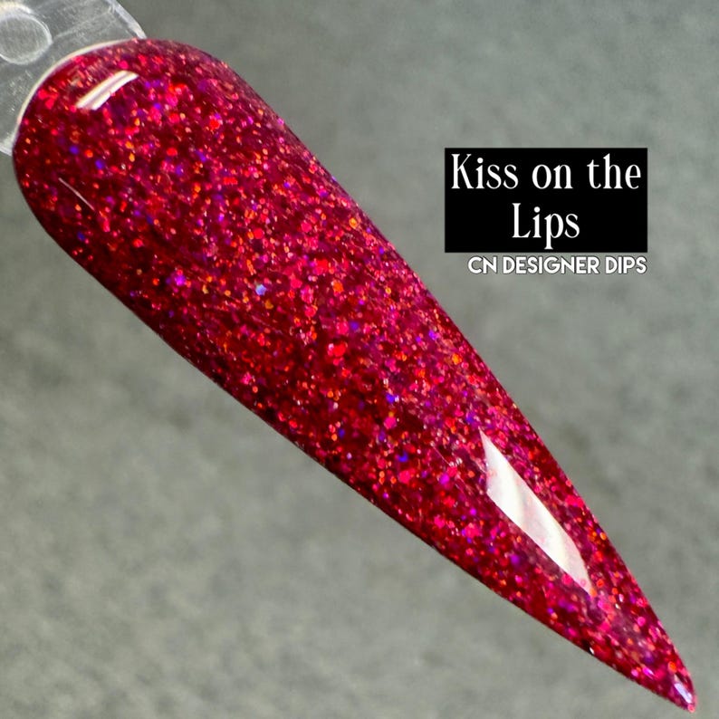 Puede incluir: Una muestra de u&ntilde;as muestra un esmalte de u&ntilde;as con purpurina roja. El esmalte es de un rojo intenso y brillante con motas de purpurina m&aacute;s clara e iridiscente. El texto "Kiss on the Lips" y "CN DESIGNER DIPS" se muestra en un rect&aacute;ngulo negro.