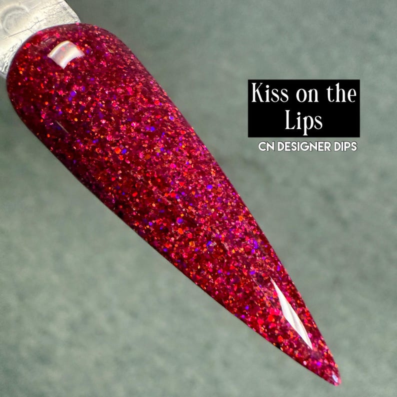 Puede incluir: Una muestra de u&ntilde;as con un vibrante acabado de purpurina roja. La u&ntilde;a tiene forma de punta. El texto "Kiss on the Lips" y "CN DESIGNER DIPS" se muestra en un rect&aacute;ngulo negro.