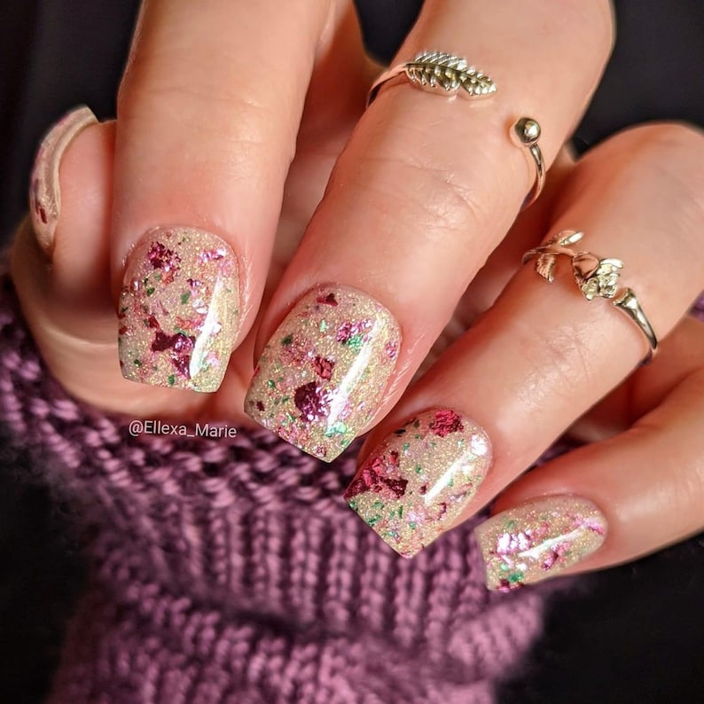 May include: Close-up of a hand with short, square-shaped nails painted with a light gold glitter nail polish with pink and green glitter accents. The nails are decorated with small, pink and green glitter flakes. The hand is wearing two silver rings, one with a leaf design and the other with a rose design. The hand is resting on a purple knitted fabric.