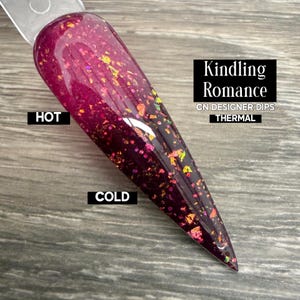 May include: A nail swatch with a thermal dip, shifting from a dark burgundy to a lighter pink with gold and iridescent glitter. The image text reads "Kindling Romance" and "Thermal". The colour changes with temperature.